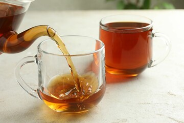 Pouring aromatic tea from teapot into cup at light textured table, closeup