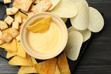 Tasty cheese dipping sauce in bowl and different snacks on dark wooden table, top view