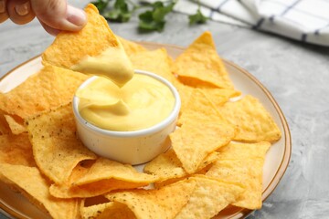 Woman dipping nacho chip into tasty cheese sauce at grey table, closeup