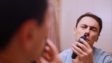 Man removing nose hair with electric trimmer in bathroom mirror
