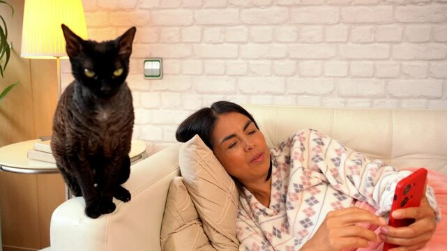 Playful interaction between a woman and her Devon Rex cat while she uses her phone on the sofa