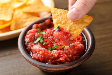 Woman dipping nacho chip into spicy salsa sauce at table, closeup