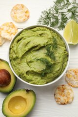 Tasty avocado dip in bowl, dill, lime and puffed rice cakes on white wooden table, flat lay