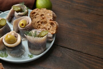 Delicious herring rolls with olives, pickles, dill and bread pieces on wooden table, closeup