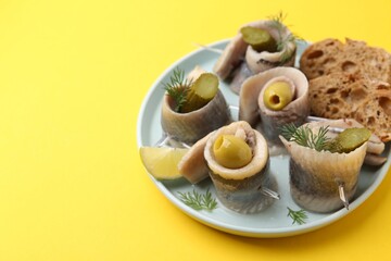 Delicious herring rolls with olives, pickles, dill and bread pieces on yellow table, closeup. Space...
