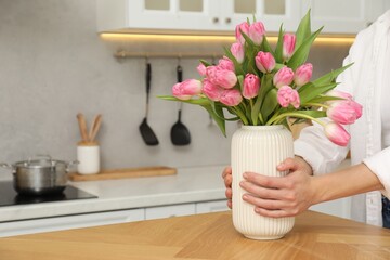 Woman holding vase with bouquet of beautiful tulips at wooden table in kitchen, closeup. Space for text