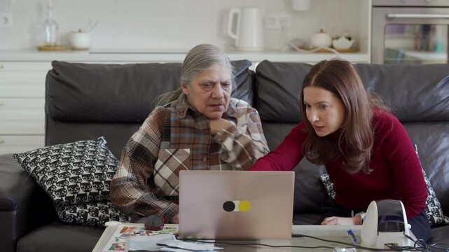 A woman is teaching her senior mother how to use a laptop for online tasks in their home, showing her something on the screen.
