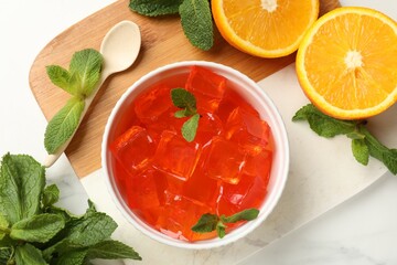 Delicious jelly cubes in bowl, mint, spoon and orange on white marble table, flat lay