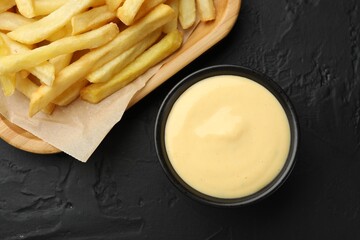 Tasty cheese sauce in bowl and French fries on black table, flat lay
