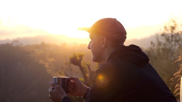 Sunrise illuminating mountain landscape while backlit hiker in cap drinking steaming beverage, embodying peaceful solitude and adventurous spirit during golden morning moment