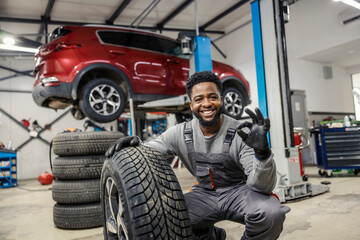 Portrait of smiling interracial auto mechanic crouching next to a tire and gesturing okay at auto...