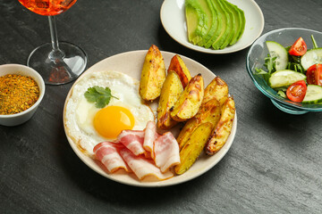 Tasty brunch. Fried egg, potato, bacon and salad on dark textured table, closeup
