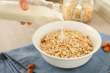 Woman pouring milk into bowl with oatmeal at table, closeup