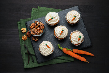 Tasty carrot muffins with cream, walnuts and fresh vegetables on black wooden table, flat lay