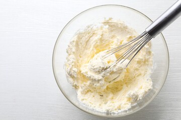 Bowl with whisk and whipped cream on white wooden table, top view