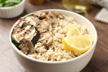 Delicious brown rice with mushrooms, zucchini and lemon slices served on wooden table, closeup