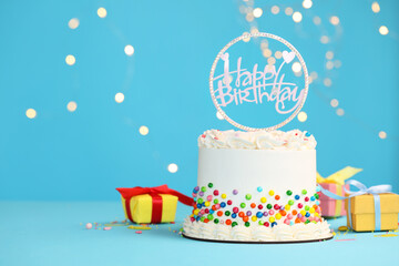 Delicious bento cake with colorful sprinkles and topper with words Happy Birthday on light blue table against blurred lights. Space for text