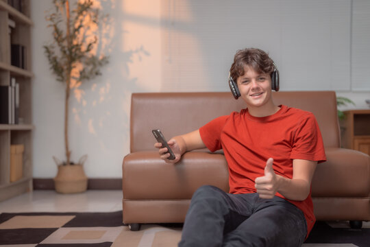 Happy teenager listening music with headphones and smartphone sitting on the floor near sofa at home, giving thumbs up, enjoying his leisure time