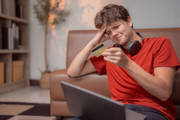 Young man looking worried while holding a credit card and trying to make an online payment with his...