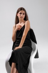 Fototapeta premium Model poses elegantly in a black dress on a minimalist chair in a bright studio setting