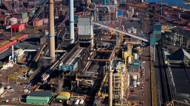 Industrial steel plant in IJmuiden, Netherlands, with large chimneys, conveyor belts, and emissions. Aerial view of a major production hub, showing both traditional and renewable energy elements.