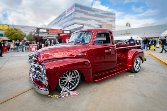 A polished red classic truck displayed at SEMA show with people in the background.