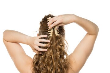 Rear view of young woman placing beige claw clip in her long wavy blonde hair, demonstrating half-up hairstyling process against white background