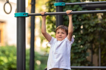 Fototapeta premium Little schoolboy, young athlete is pulling himself up on a sport horizontal bar on outdoor playground on sunny day. Active childhood, acrobat, workout on backyard, summer camp concept