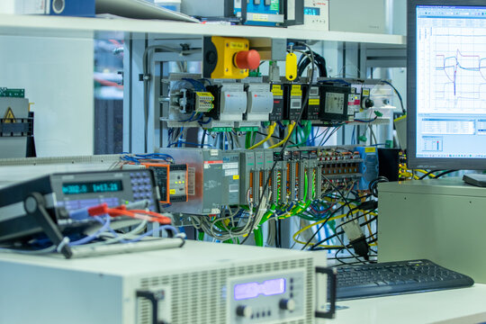 Wide view of industrial automation test bench with electronic measuring devices, power supply, PLC modules, and monitoring software on screen in laboratory setting