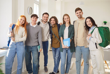 Fototapeta premium Group portrait of cheerful young students standing together in modern classroom and smiling at camera. Happy friends with backpacks and notebooks hugging each other, showing unity and friendship.