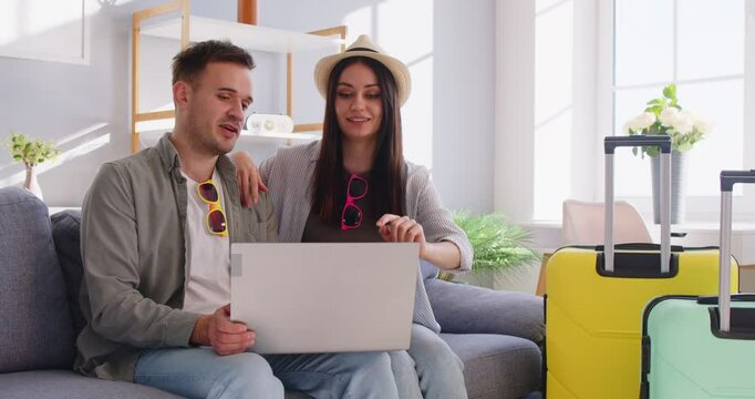 Family husband and wife choosing tour for vacation online, booking hotel room, discussing something while concentrated at screen, posing on sofa near packed suitcases, going on leisure trip