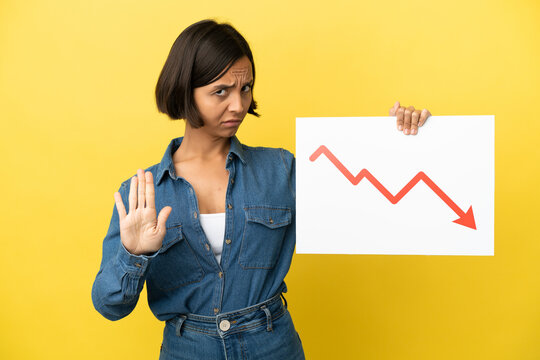 Young mixed race woman isolated on yellow background holding a sign with a decreasing statistics arrow symbol and doing stop sign