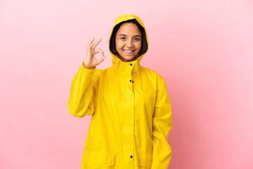 Young latin woman wearing a rainproof coat over isolated background showing ok sign with fingers