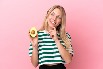 Young caucasian woman holding an avocado isolated on pink background looking up while smiling