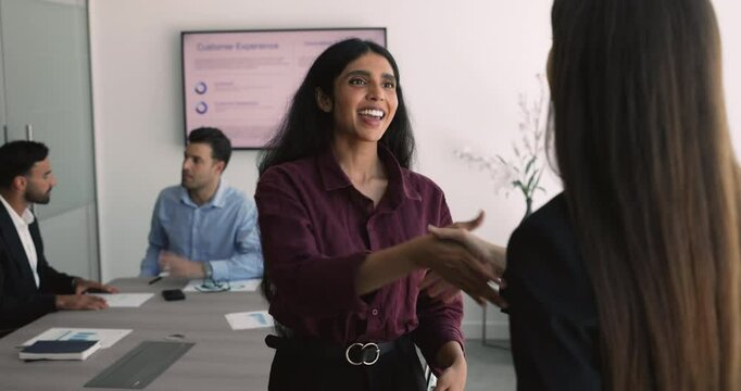 Two multiethnic businesswomen talking, shaking hands, accomplish peasant professional conversation, diverse colleagues lead discussion seated at conference desk in background. Making deal, business