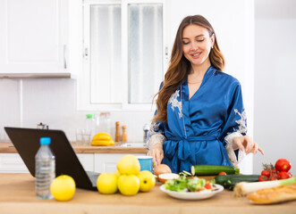 Young woman in blue robe cooking and watching TV series on laptop