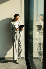 Professional Asian woman smiles while using a tablet near a large window during daytime
