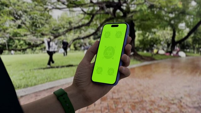 GREEN SCREEN CHROMA KEY Person holding a smartphone with a green screen in an outdoor park setting, surrounded by trees, grass, and people enjoying leisure