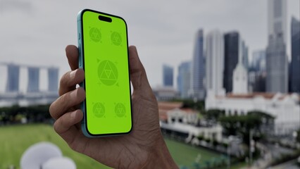 GREEN SCREEN CHROMA KEY Person holding a smartphone with a green screen outdoors, overlooking a modern urban skyline and grassy area on a cloudy day