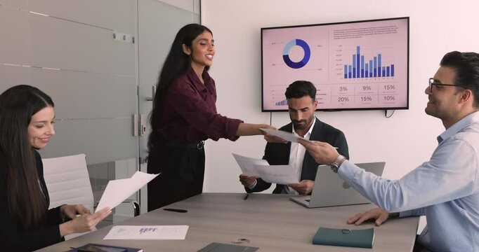 Indian female project manager offering plan, management strategy to multinational professional team, handing report papers, sharing document or contract for signing during group meeting or negotiation