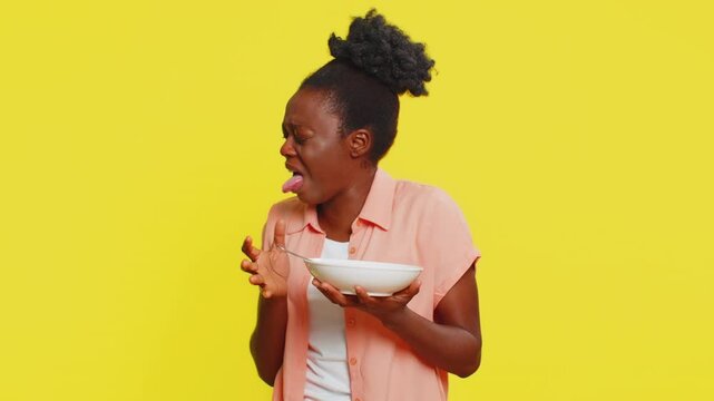African American woman holds spoon and bowl, grimacing after tasting taking a sip overly salty, spicy or hot soup. Black girl on yellow background expresses disappointment, dissatisfaction with meal.