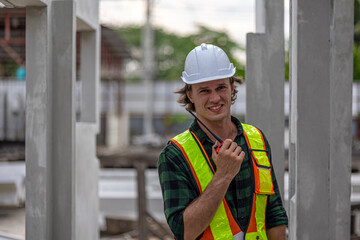 Male civil engineer with walkie talkie and laptop stands next to ready made wall or precast boards conducting a quality check in a factory. Engineer in safety suit working in a precast factory.