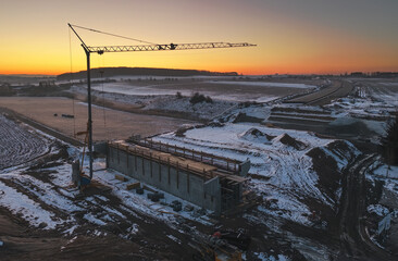Construction of a bridge on the highway D35 during a snowy winter morning. The silhouette of a crane against the morning colored sky at sunrise.