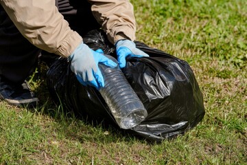 a young male person wearing gloves, picking up a plastic bottle and other garbage from the grass in a park