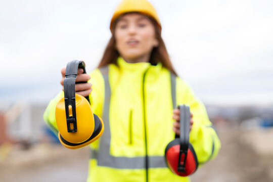 Female Worker demonstrates safety ear protection on construction site while wearing safety vest and hard hat