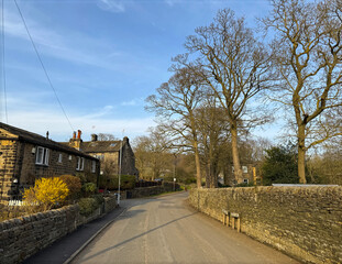 A winters day on Esholt Lane, as it winds through, Esholt village, bordered by rustic stone houses and leafless trees under a clear blue sky. The warm sunlight highlights the peaceful scene.