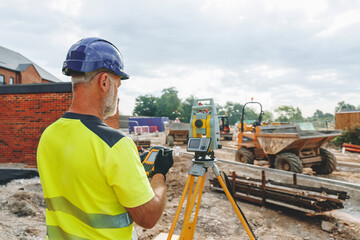Builder engineer operating surveying equipment on a building site with machinery in the background