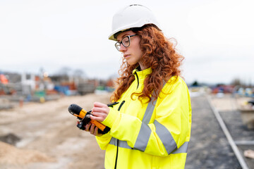 Female builder checks rugged mobile handheld device controller while wearing safety jacket and maintaining focus on her task © Iryna