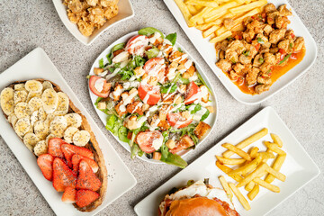 A top view of diverse dishes including salad, fries, toast with fruits, fried chicken, and a burger, all served on white plates over a neutral background.
