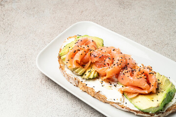 A delicious toast topped with smoked salmon, fresh avocado, cream cheese, and sesame seeds, served on a white plate over a textured neutral background.